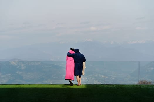Back view of anonymous couple in robes looking away while standing closely and hugging on green lawn and admiring view of mountains and hazy nature in daylight