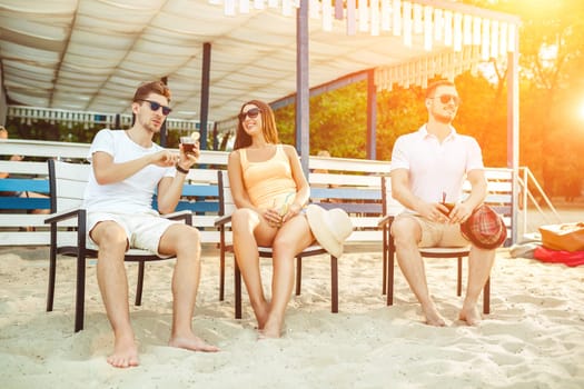 Lifestyle Young people enjoying summer vacation sunbathing drinking at beach bar. Two guys and a young girl sit on the terrace of the summer bar and enjoy the conversation Sun flare