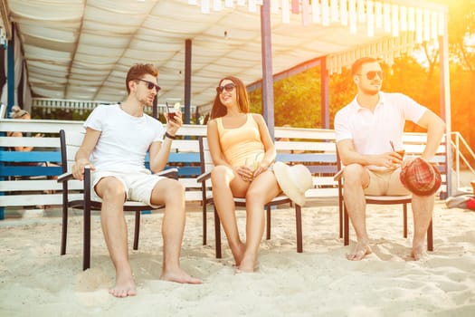 Lifestyle Young people enjoying summer vacation sunbathing drinking at beach bar. Two guys and a young girl sit on the terrace of the summer bar and enjoy the conversation Sun flare