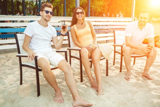 Lifestyle Young people enjoying summer vacation sunbathing drinking at beach bar. Two guys and a young girl sit on the terrace of the summer bar and enjoy the conversation Sun flare