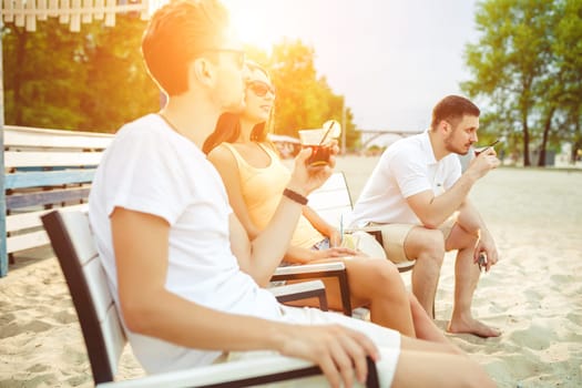 Lifestyle Young people enjoying summer vacation sunbathing drinking at beach bar. Two guys and a young girl sit on the terrace of the summer bar and enjoy the conversation Sun flare