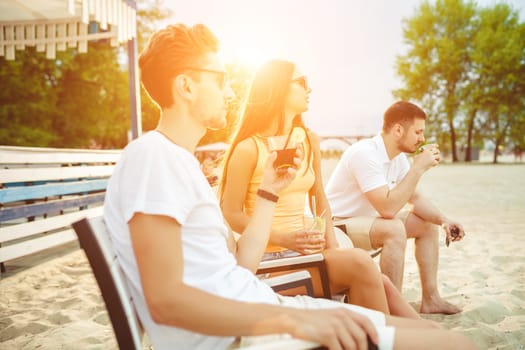 Lifestyle Young people enjoying summer vacation sunbathing drinking at beach bar. Two guys and a young girl sit on the terrace of the summer bar and enjoy the conversation Sun flare