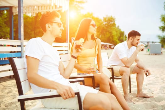 Lifestyle Young people enjoying summer vacation sunbathing drinking at beach bar. Two guys and a young girl sit on the terrace of the summer bar and enjoy the conversation Sun flare