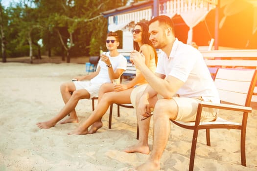 Lifestyle Young people enjoying summer vacation sunbathing drinking at beach bar. Two guys and a young girl sit on the terrace of the summer bar and enjoy the conversation Sun flare