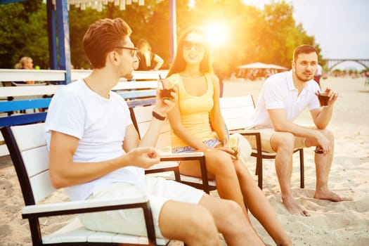 Lifestyle Young people enjoying summer vacation sunbathing drinking at beach bar. Two guys and a young girl sit on the terrace of the summer bar and enjoy the conversation Sun flare