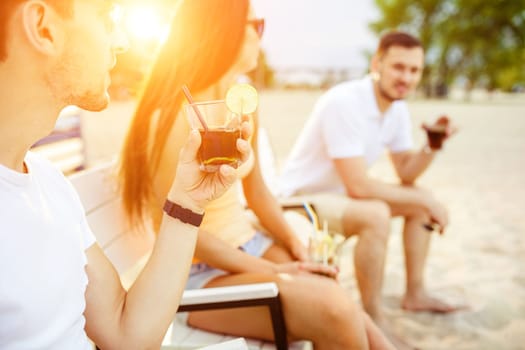 Lifestyle Young people enjoying summer vacation sunbathing drinking at beach bar. Two guys and a young girl sit on the terrace of the summer bar and enjoy the conversation Sun flare