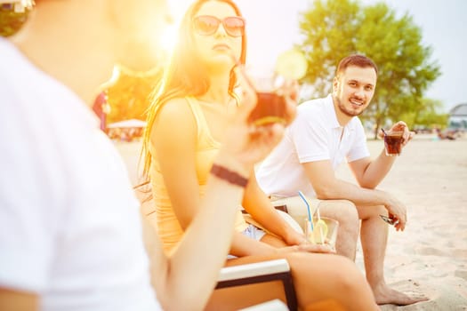 Lifestyle Young people enjoying summer vacation sunbathing drinking at beach bar. Two guys and a young girl sit on the terrace of the summer bar and enjoy the conversation Sun flare