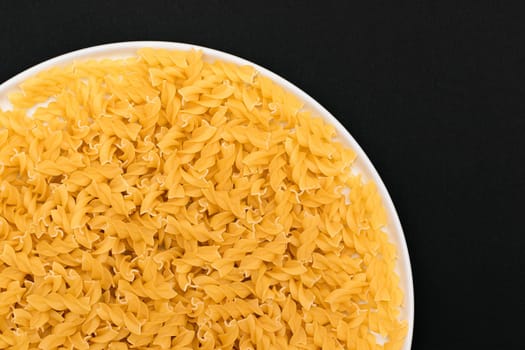 Uncooked Fusilli Pasta Lying on White Plate on Black Background. Raw and Dry Macaroni. Unhealthy and Fat Food - Top View