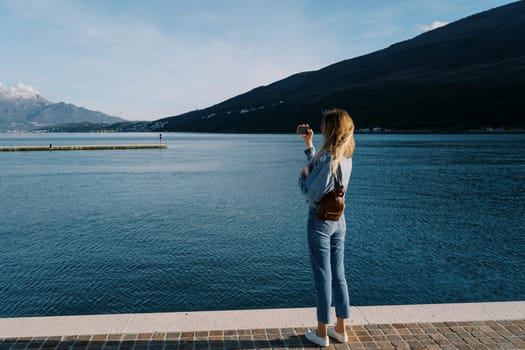Woman takes pictures of the sea and mountains with a smartphone while standing on the pier. Back view. High quality photo