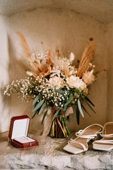Wedding rings in a box stand in a niche in a stone wall near bridal bouquet and shoes. High quality photo