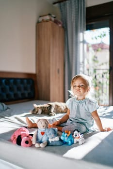 Little girl sits on a bed in front of a row of plush toys. High quality photo