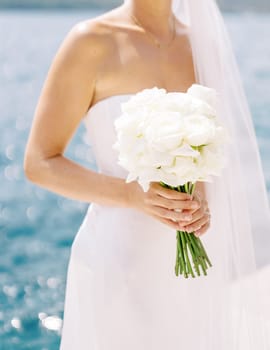 Bouquet of flowers in the hands of a bride standing on the seashore. Cropped. Faceless. High quality photo