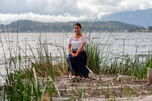 pretty indigenous woman from Ecuador sitting on a dock surrounded by sea plants on the shore of a large lake. High quality photo