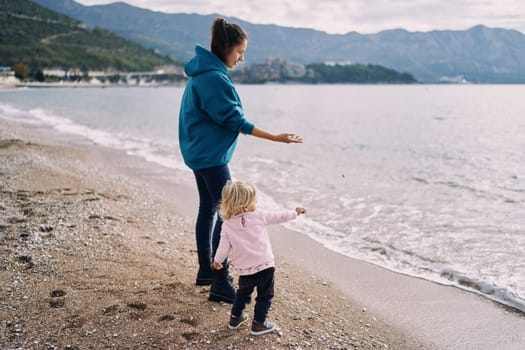 Mom and her little daughter are standing on the seashore and throwing pebbles into the water. High quality photo