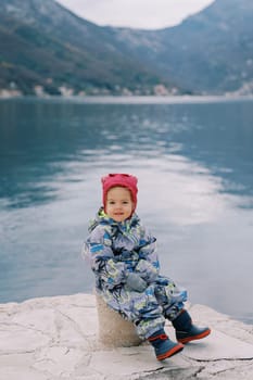 Little smiling girl sitting on a bollard on the pier. High quality photo
