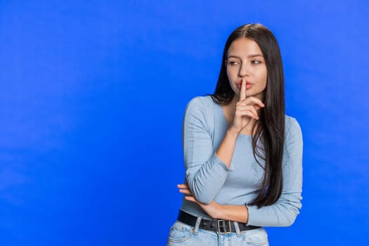 Shh be quiet please. Young woman presses index finger to lips makes silence gesture sign do not tells secret, stop gossip. Pretty girl isolated alone on blue studio background. Copy-space for text