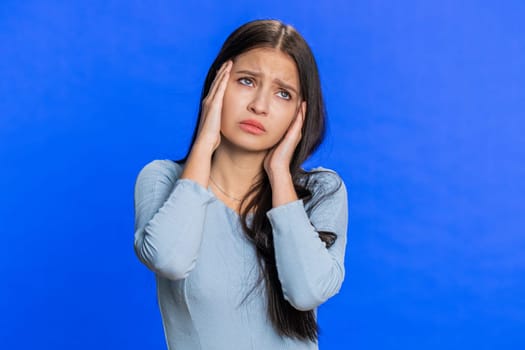 Attractive exhausted sad young woman rubbing temples to cure headache problem, suffering from tension, migraine, stress, grimacing in pain, high blood pressure. Girl isolated on blue wall background