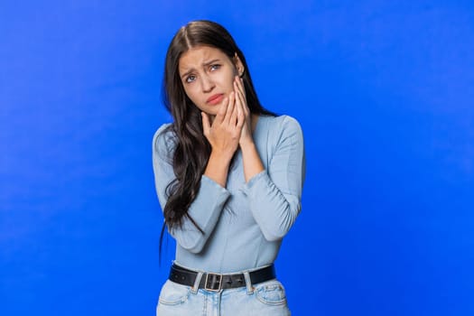 Dental problems. Young woman touching cheek, closing eyes with expression of terrible suffer from painful toothache, sensitive teeth, cavities. Pretty sad girl isolated on blue studio background