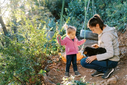Little girl stands near a swing and watches her mom petting a puppy on her lap in the forest. High quality photo