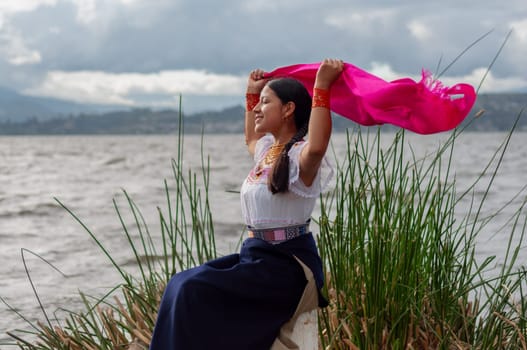indigenous girl sitting on the shore of a lake feeling the breeze while holding a fuchsia veil. High quality photo