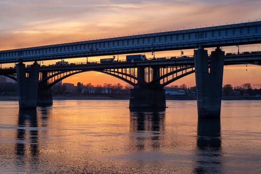 Beautiful view of the bridge over which cars drive at sunset. A river flows under the bridge, reflecting the sunset rays. Bridge in the city of Novosibirsk, Russia