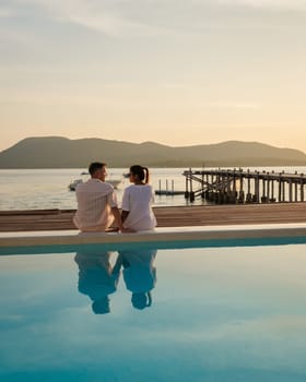 A couple of European men and an Asian woman on vacation in Thailand sitting on the edge of a pool by the ocean, Wooden pier in the ocean during sunset in Samaesan Thailand