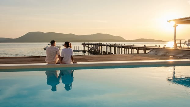 A couple of European men and an Asian woman on vacation in Thailand sitting on the edge of a pool by the ocean, couple watching the sunset in the evening in the ocean in Samaesan Thailand