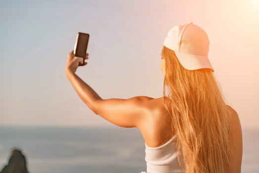 Selfie woman sea. The picture depicts a woman in a cap and tank top, taking a selfie shot with her mobile phone, showcasing her happy and carefree vacation mood against the beautiful sea backgroun.