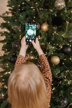 A little blonde girl photographs balloons on a Christmas tree in a festive interior decorated in a New Year's style. The concept of a merry Christmas