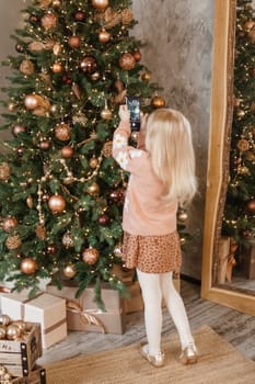 A little blonde girl photographs balloons on a Christmas tree in a festive interior decorated in a New Year's style. The concept of a merry Christmas