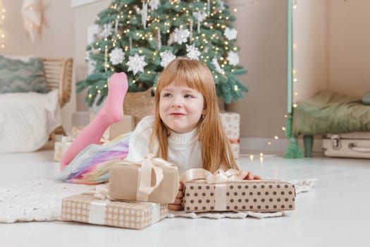 A little red-haired girl lies next to Christmas gifts in a craft package. Happy Christmas concept