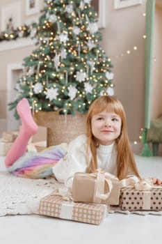 A little red-haired girl lies next to Christmas gifts in a craft package. Happy Christmas concept