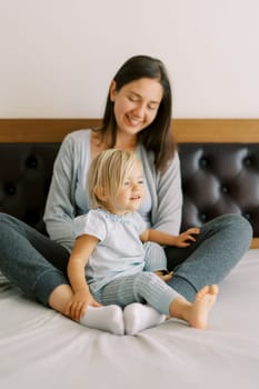 Smiling mother looking at little girl sitting on bed with her. High quality photo