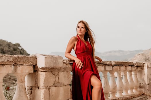 Side view a Young beautiful sensual woman in a red long dress posing on a volcanic rock high above the sea during sunset. Girl on the nature on overcast sky background. Fashion photo