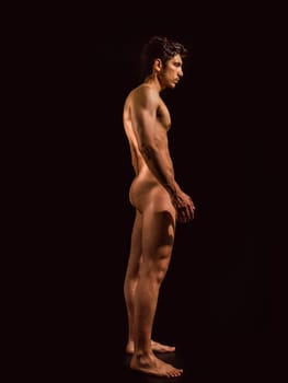 Totally naked athletic handsome young man, seen from the back, showing buttocks, looking straight in a muscular tension pose, on dark background in studio. Portrait of naked handsome young man with languishing look covering crotch with a hand, black background