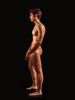 Totally naked athletic handsome young man, seen from the back, showing buttocks, looking straight in a muscular tension pose, on dark background in studio. Portrait of naked handsome young man with languishing look covering crotch with a hand, black background