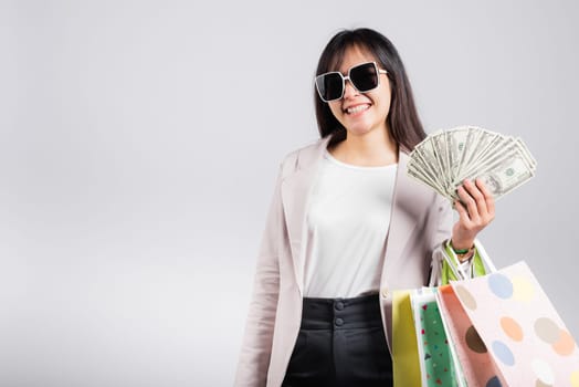 Woman with glasses confident shopper smile holding online shopping bags multicolor and dollar money banknotes on hand, excited happy Asian young female person studio shot isolated on white background