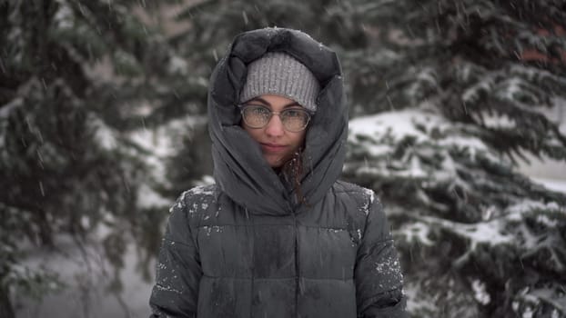 A young woman stands against a background of fir trees under heavy snowfall in winter. A girl in glasses and a down jacket with a hood stands and looks at the camera. Closeup. 4k