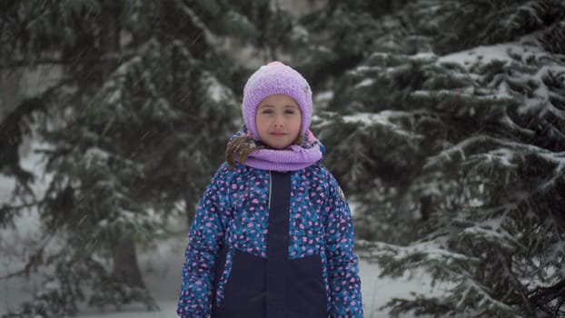 A girl stands against a background of fir trees under snowfall and looks at the camera. A girl in a jacket and hat stands under heavy snowfall. 4k