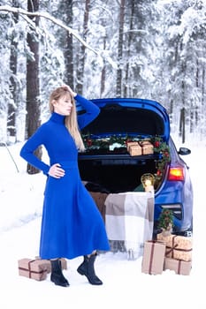 Woman in winter snowy forest in blue dress next to blue car decorated with Christmas decor. Christmas and winter holidays concept