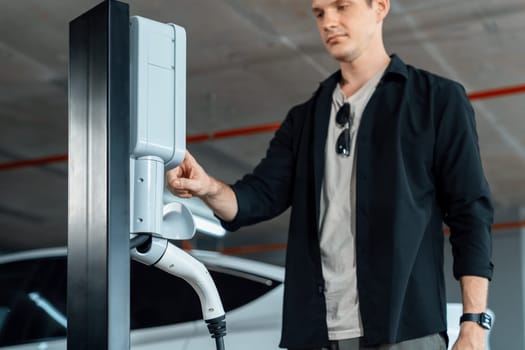 Young man travel with EV electric car to shopping center parking lot charging in downtown city showing urban sustainability lifestyle by green clean rechargeable energy of electric vehicle innards