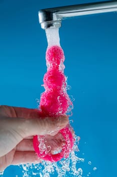 Woman holding pink anal beads under running water on blue background. Sex toy hygiene concept