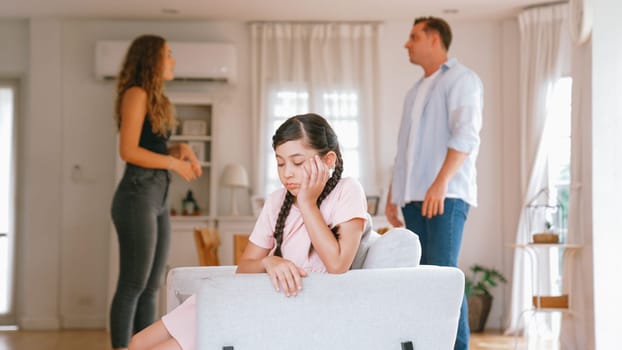 Annoyed and unhappy young girl sitting on sofa trapped in middle of tension by her parent argument in living room. Unhealthy domestic lifestyle and traumatic childhood develop to depression Synchronos