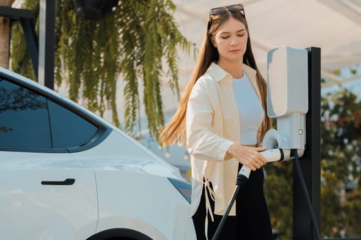 Young woman recharge her EV electric vehicle at green city park parking lot. Urban sustainability lifestyle for environmental friendly EV car with battery charging station. Expedient