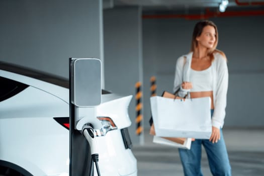 Young woman travel with EV electric car to shopping center parking lot charging in downtown city showing urban sustainability lifestyle by green clean rechargeable energy of electric vehicle innards