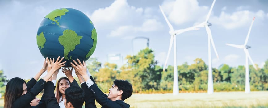 Earth day concept with big Earth globe held by asian business people team promoting environmental awareness using clean sustainable and renewable energy with wind turbine for greener future. Gyre