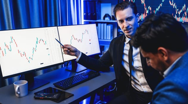 Two stock exchange traders discussing on dynamic investment graph, trending of high profit in currency stock. Showing financial benefit data on monitor screen in neon light at workplace. Sellable.