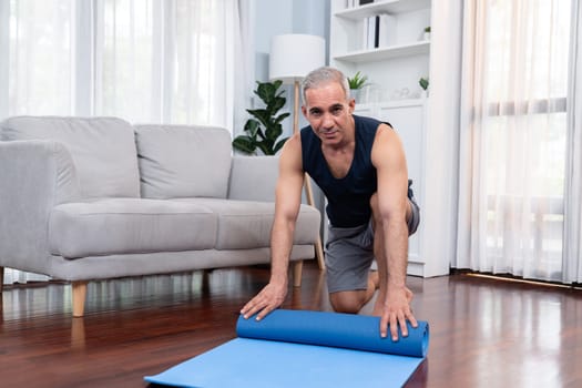 Active and sporty senior man preparing, rolling fitness exercising mat on living room floor at home. Home exercise as concept of healthy fit body lifestyle after retirement. Clout