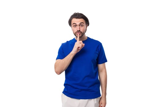 young brutal caucasian brunette man with a beard in a blue t-shirt keeps the secret of business development on a white background with copy space.