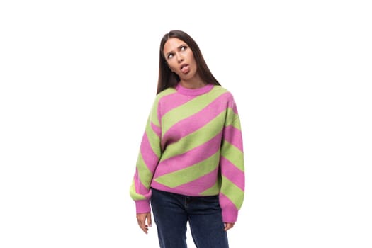young slender caucasian woman with straight black hair dressed in a striped pink pullover shows her tongue on a white background.